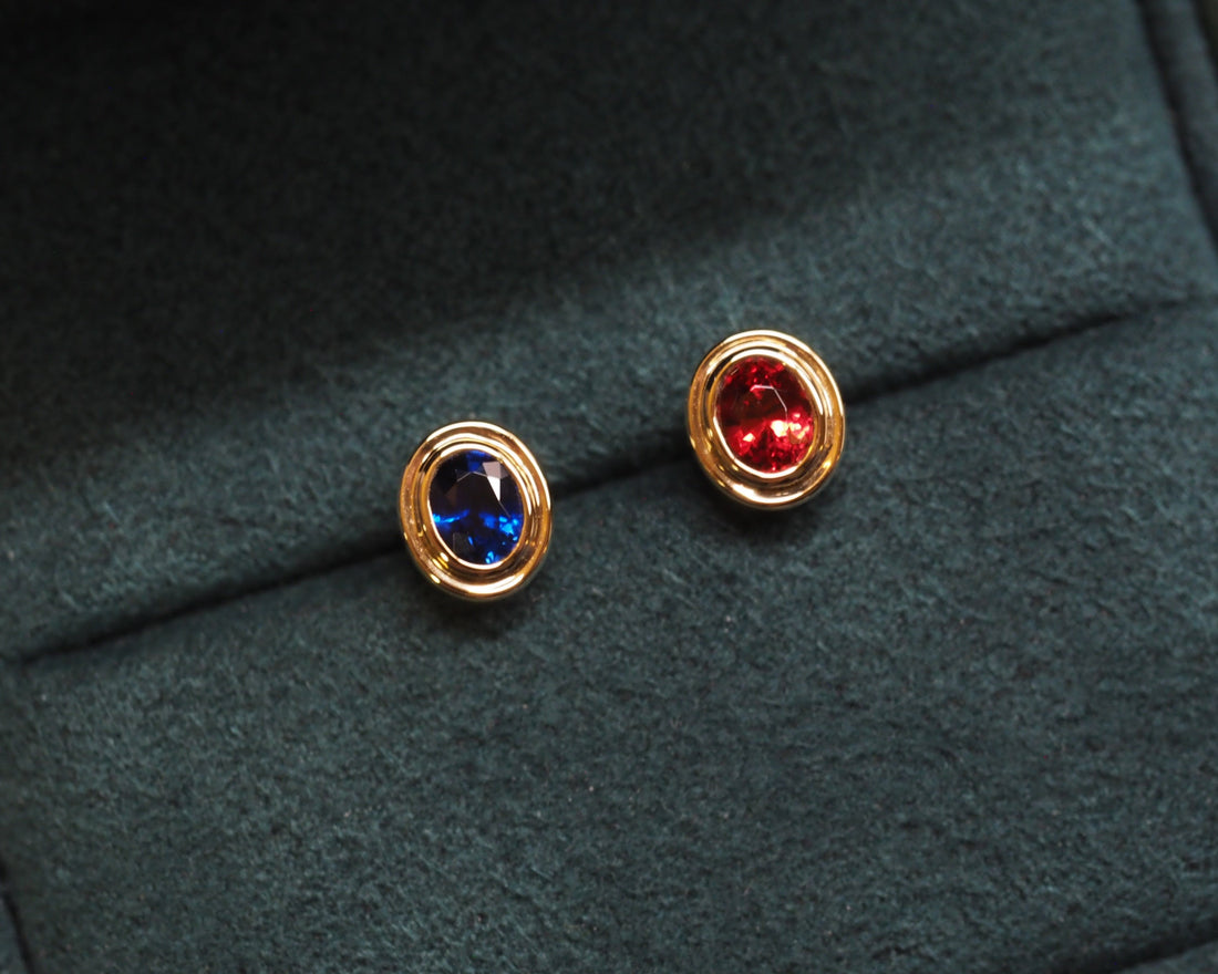Carter Mismatch Studs featuring one royal blue sapphire and a vanadium spinel, placing in front of a dark green velvet background. 