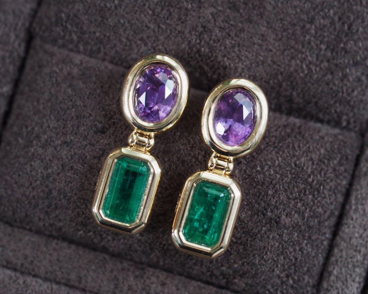 A pair of gold drop earrings featuring oval purple gemstones at the top and rectangular green emerald-cut gemstones at the bottom, displayed on a dark textured velvet surface.
