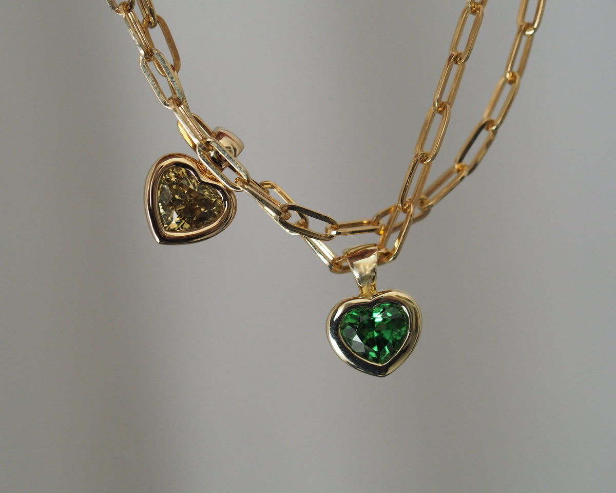 A gold chain necklace featuring two heart-shaped charms, one with a clear faceted gemstone and the other with a green faceted gemstone, both set in gold.