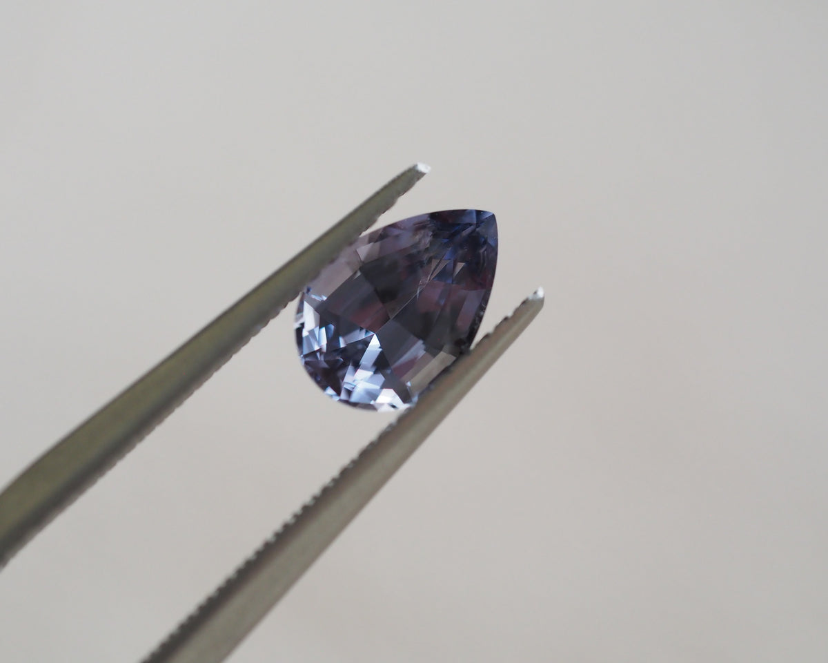 A teardrop-shaped faceted gemstone in a deep periwinkle or lavender-blue shade, held between metal tweezers against a plain light gray background.