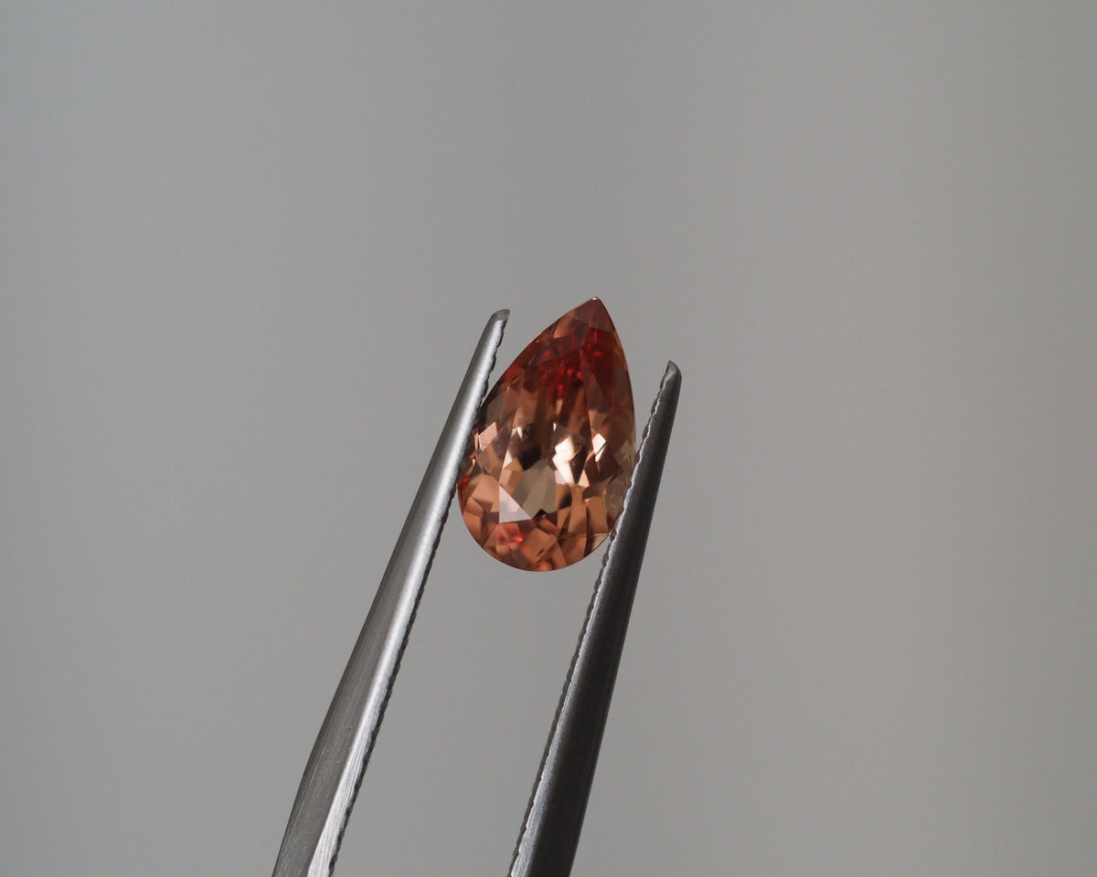 A teardrop-shaped peach-colored gemstone held between silver metal tweezers against a plain gray background.
