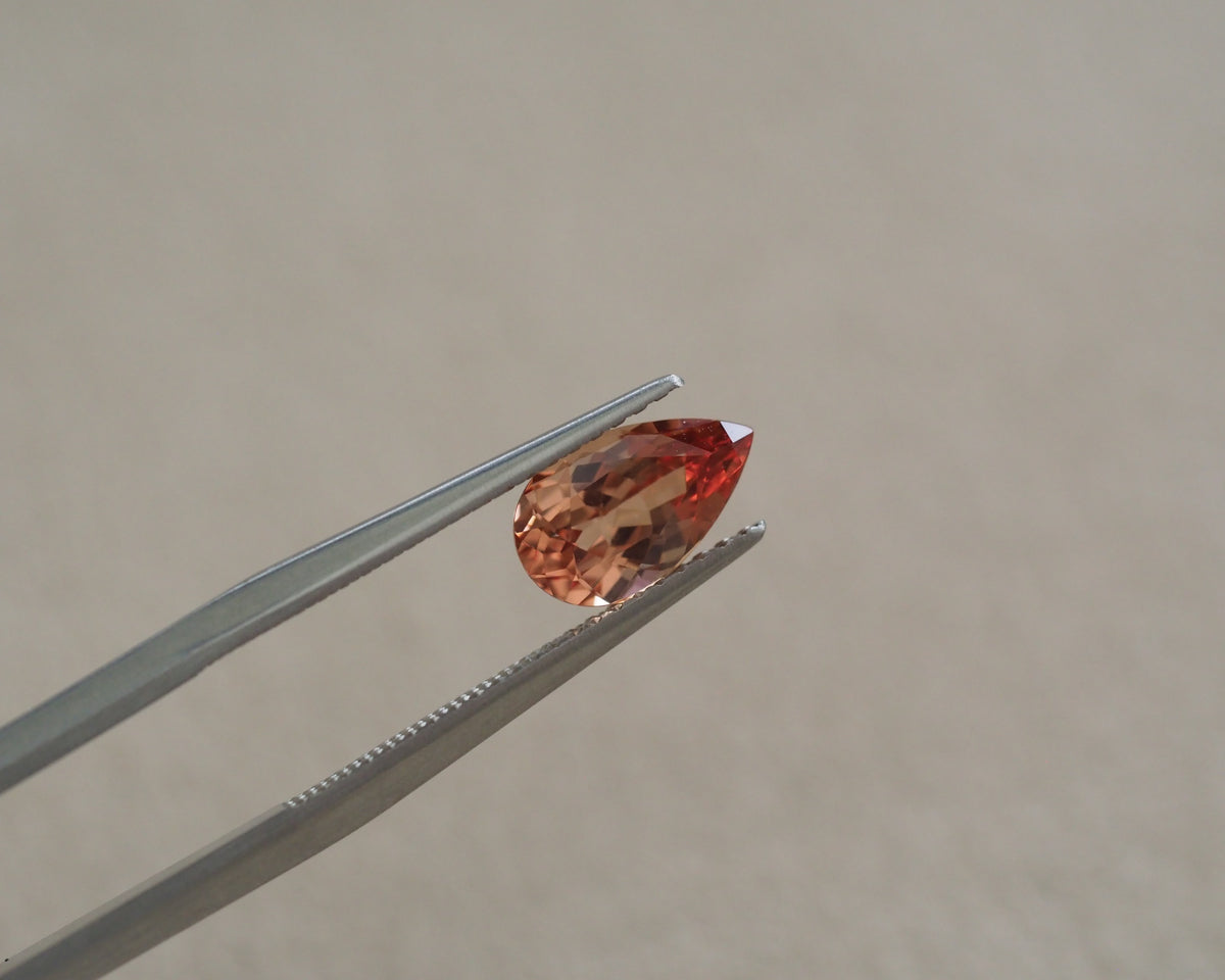 A faceted pear-cut orange gemstone held between silver metal tweezers against a plain beige background.