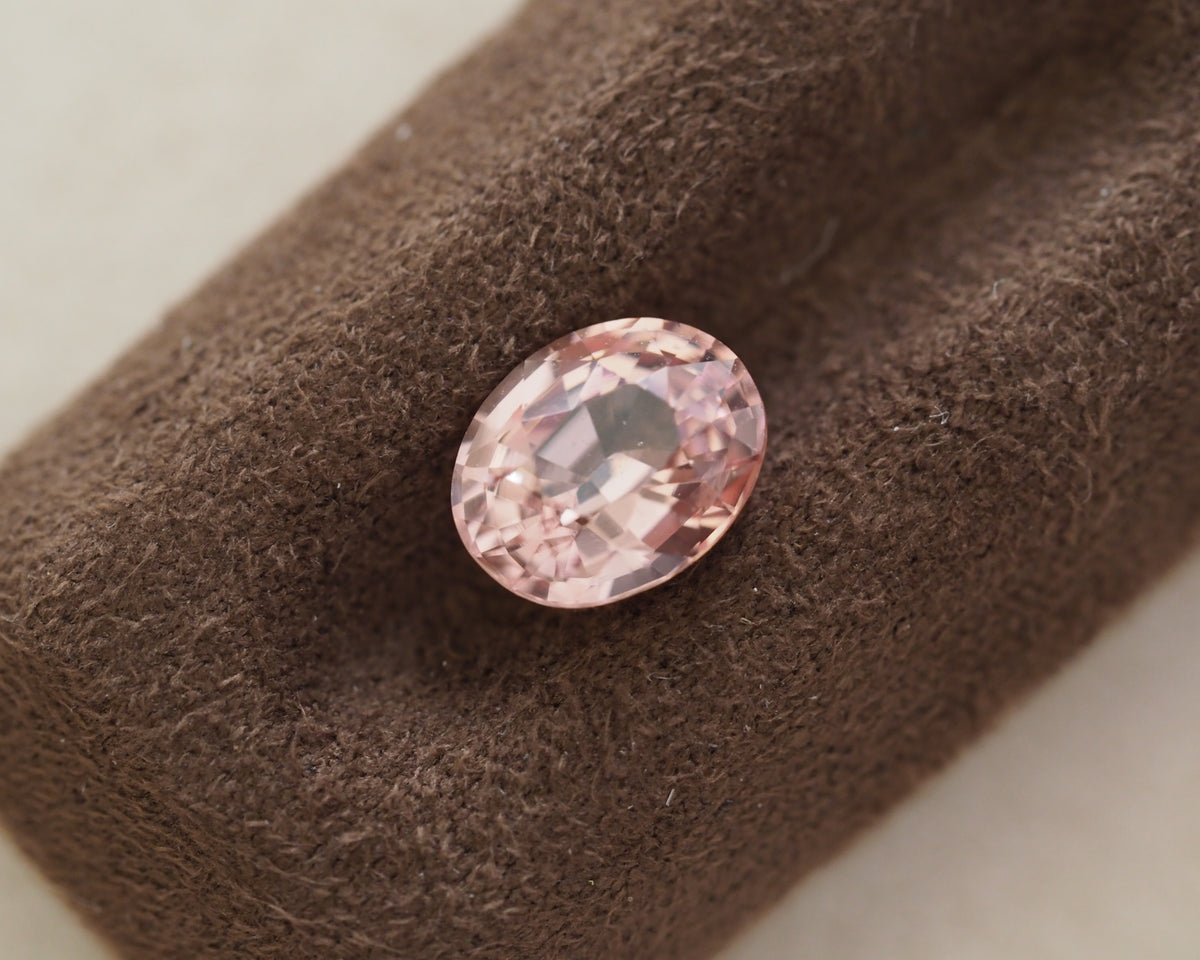 An oval-cut pink gemstone displayed on a textured brown velvet ring stand against a light neutral background.