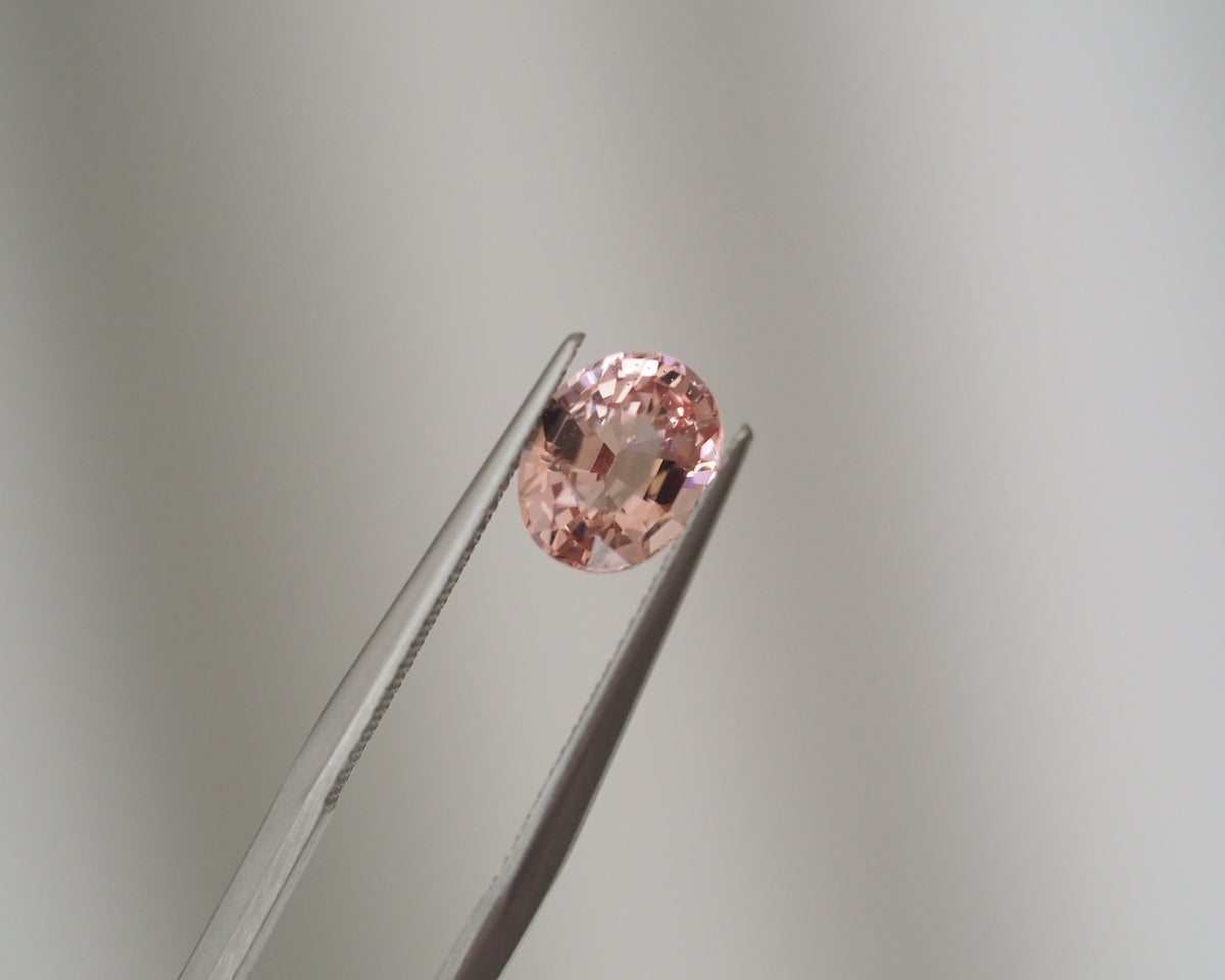 A small oval-cut pink gemstone held between metal tweezers against a soft light gray background.