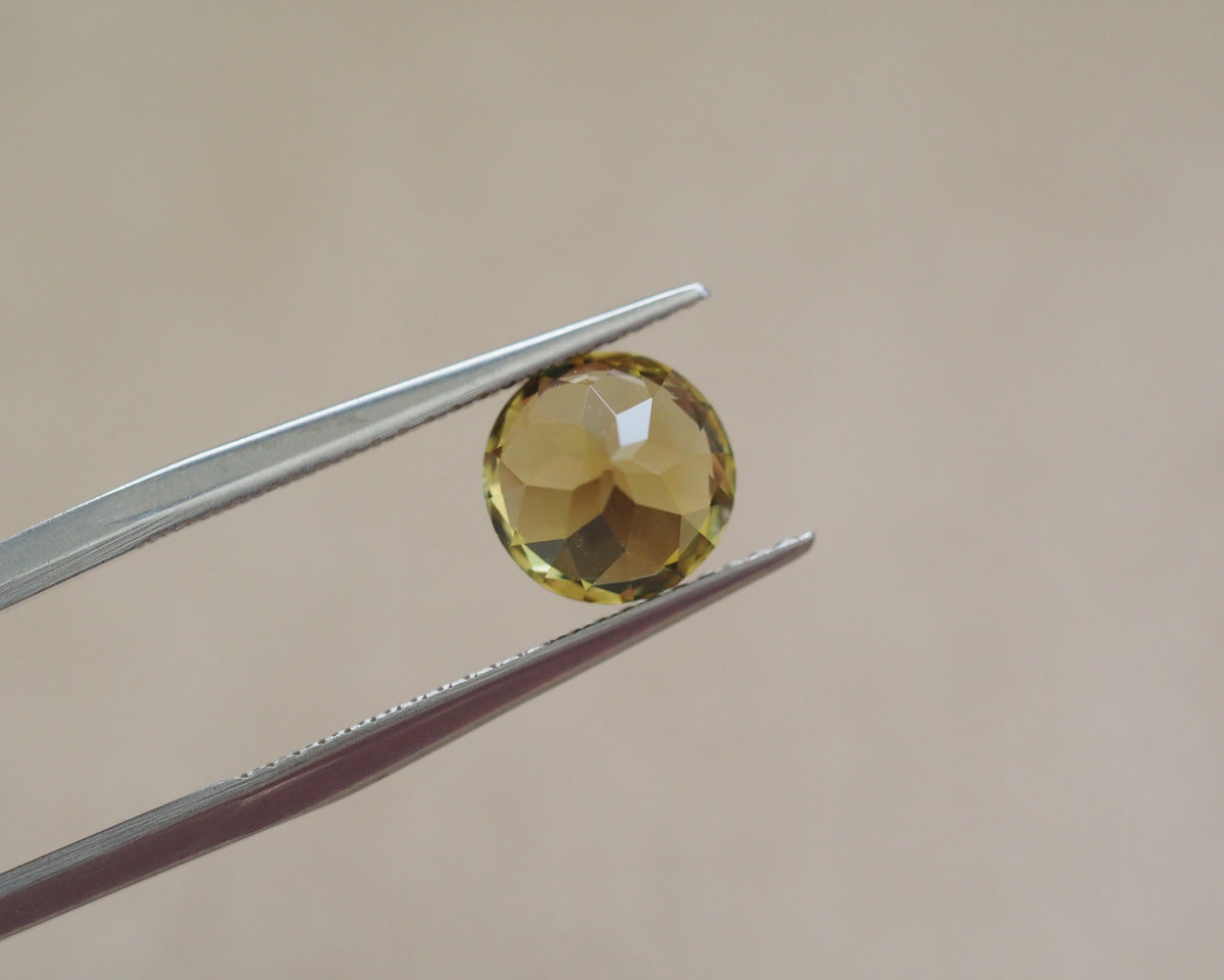 A round faceted yellow gemstone held between silver metal tweezers against a soft beige background.