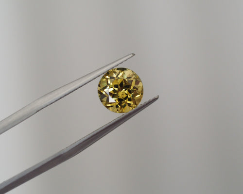 A round yellow gemstone held between metal tweezers against a plain light gray background.