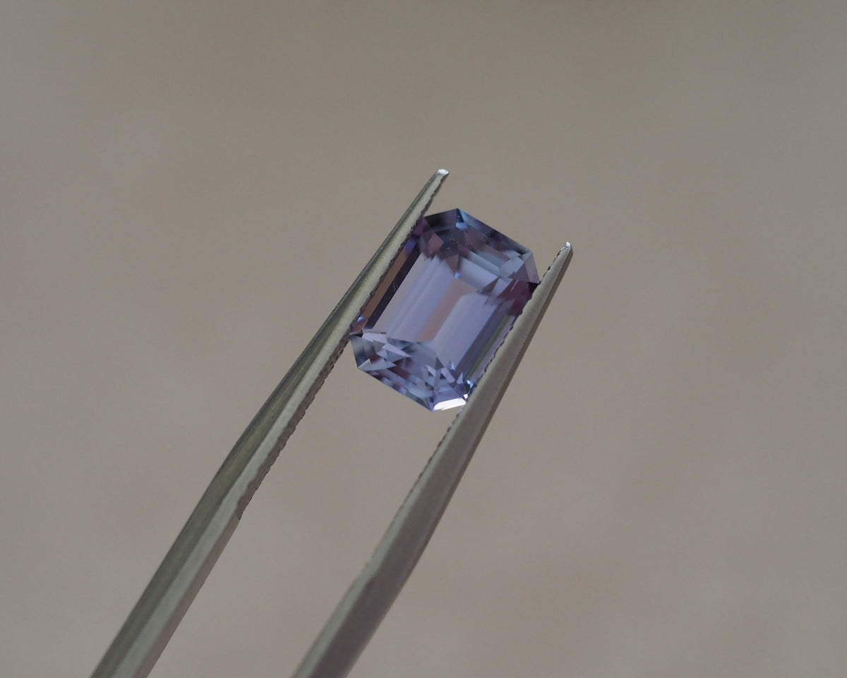 A rectangular emerald-cut lavender gemstone held in silver tweezers against a plain beige background.