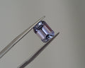 A loose rectangular emerald-cut lavender gemstone held between metal tweezers against a plain light gray background.