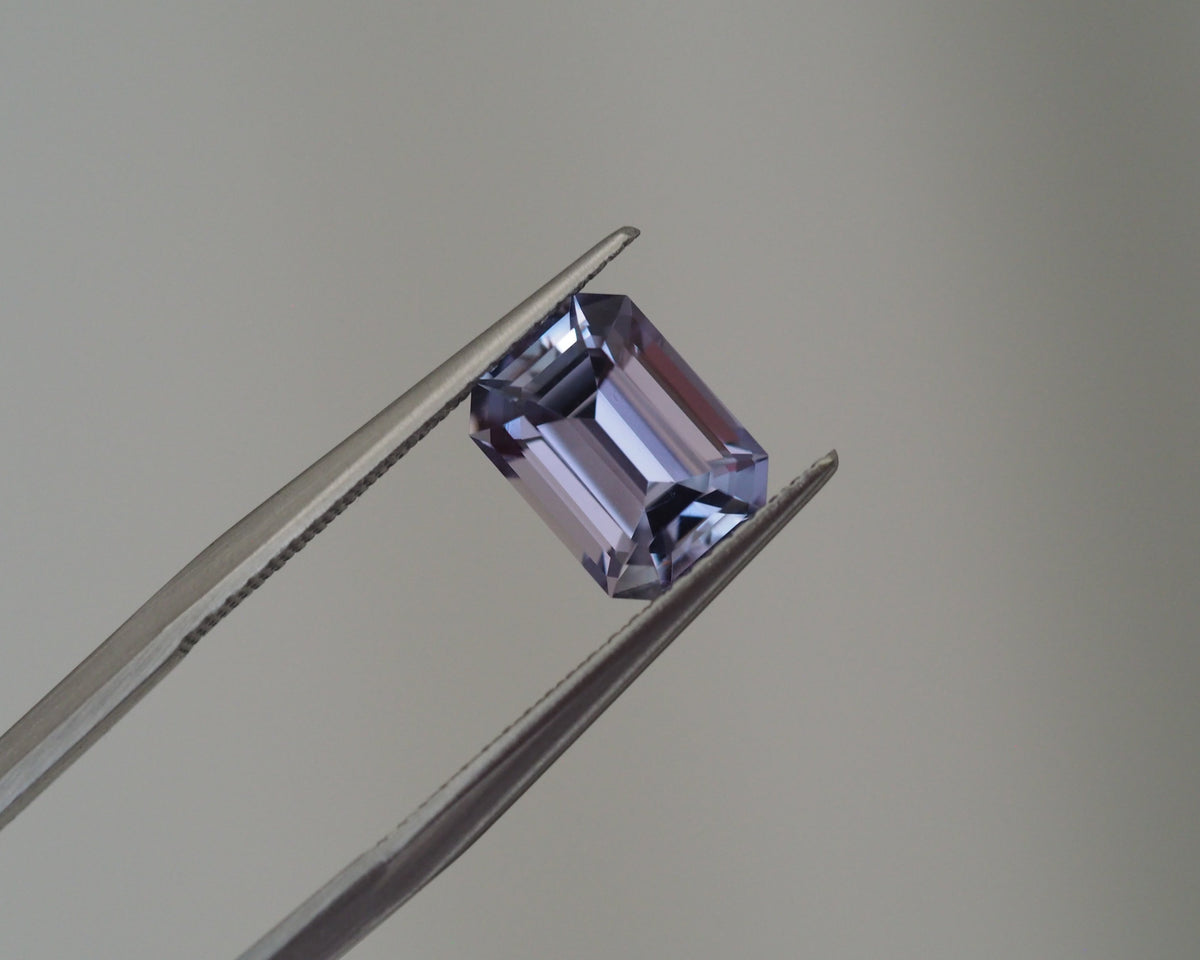 A loose rectangular emerald-cut lavender gemstone held between metal tweezers against a plain light gray background.