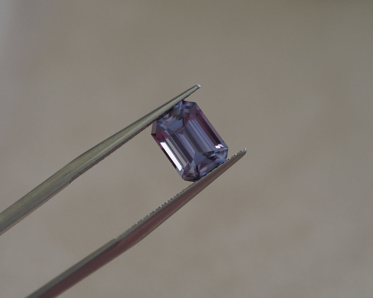 A rectangular emerald-cut purple gemstone held between metal tweezers against a soft beige background.