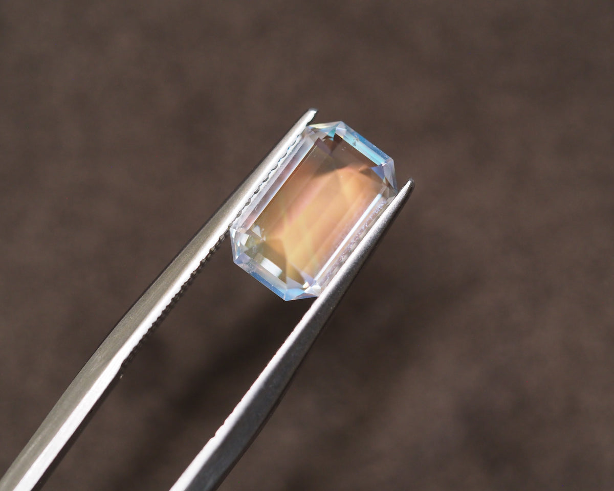 A rectangular emerald-cut clear gemstone with a warm peach-orange internal color, held in the jaws of silver metal tweezers against a dark brown textured background.