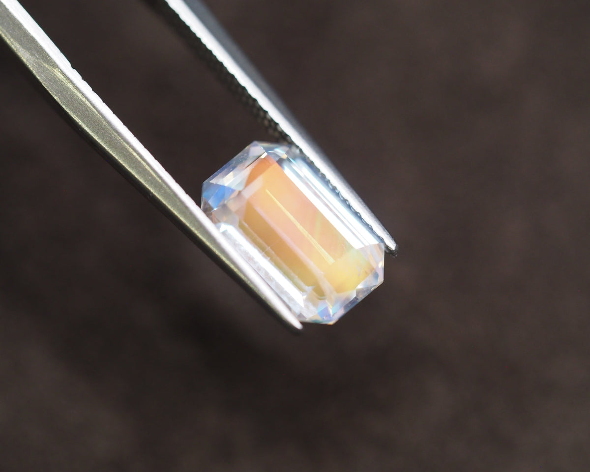 A small rectangular faceted gemstone with an iridescent peach and blue sheen, held between metal tweezers against a dark brown background.