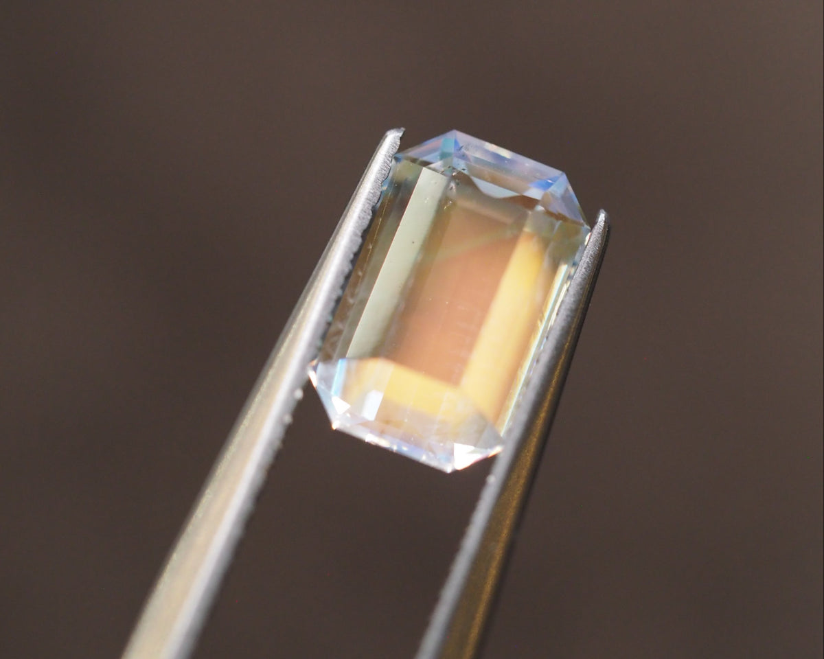 A rectangular clear gemstone with an iridescent aurora borealis effect, held in silver metal tweezers against a dark brown background.
