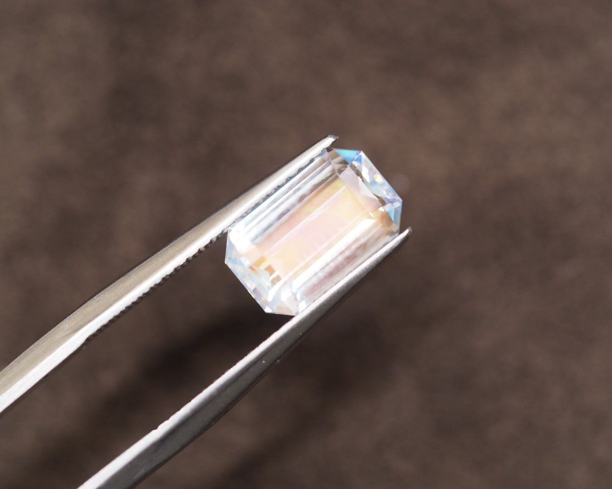 A rectangular clear gemstone with a soft yellow center and blue corner flashes, held in silver tweezers against a dark brown textured background.