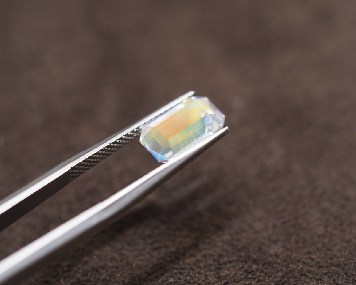 A rectangular iridescent gemstone held between silver tweezers against a dark brown textured background.