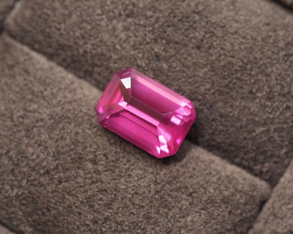 A bright pink rectangular emerald-cut Rhodolite resting on a textured brown felt surface.