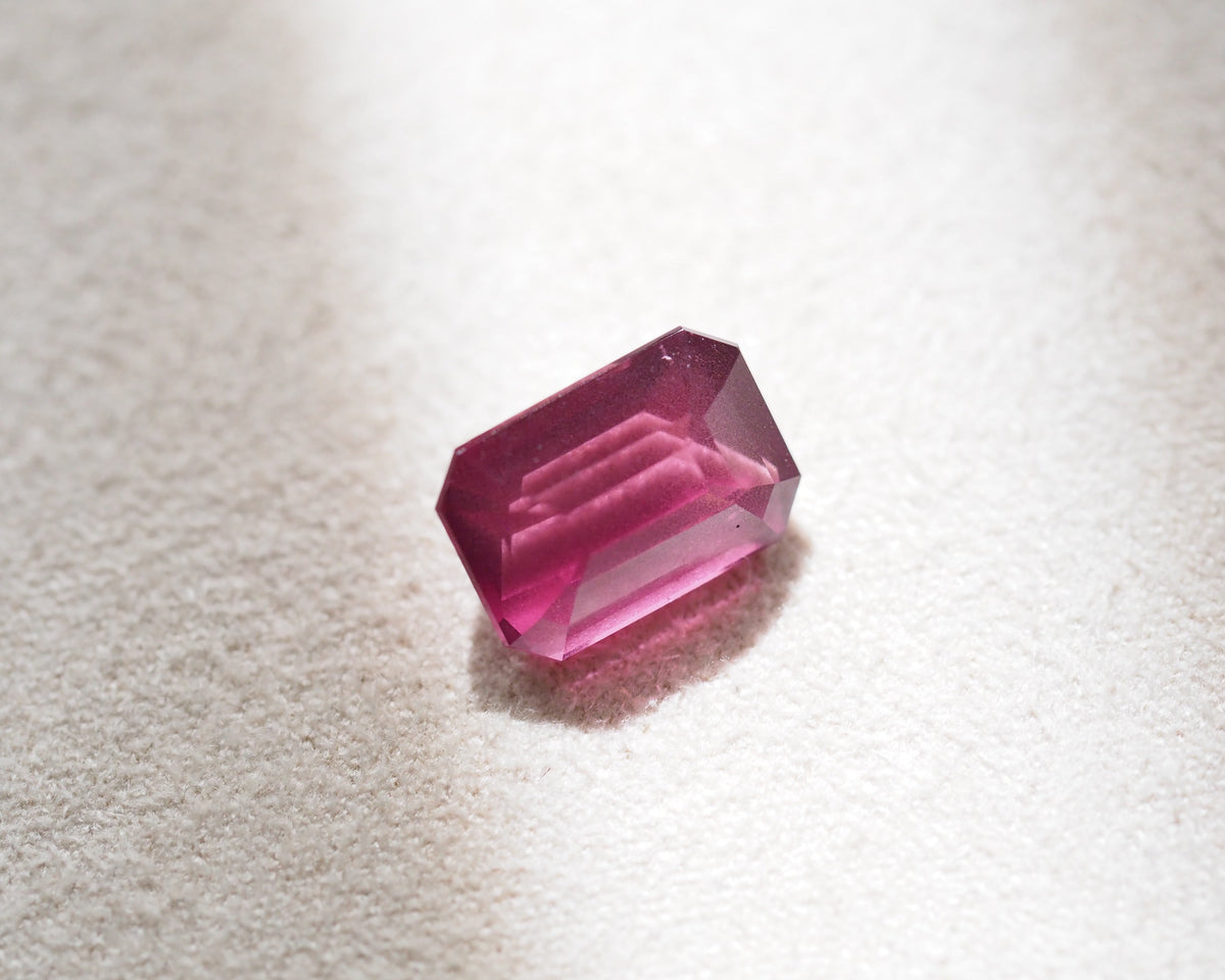 A single rectangular emerald-cut pink rhodolite with faceted edges, resting on a textured off-white surface with a soft shadow.