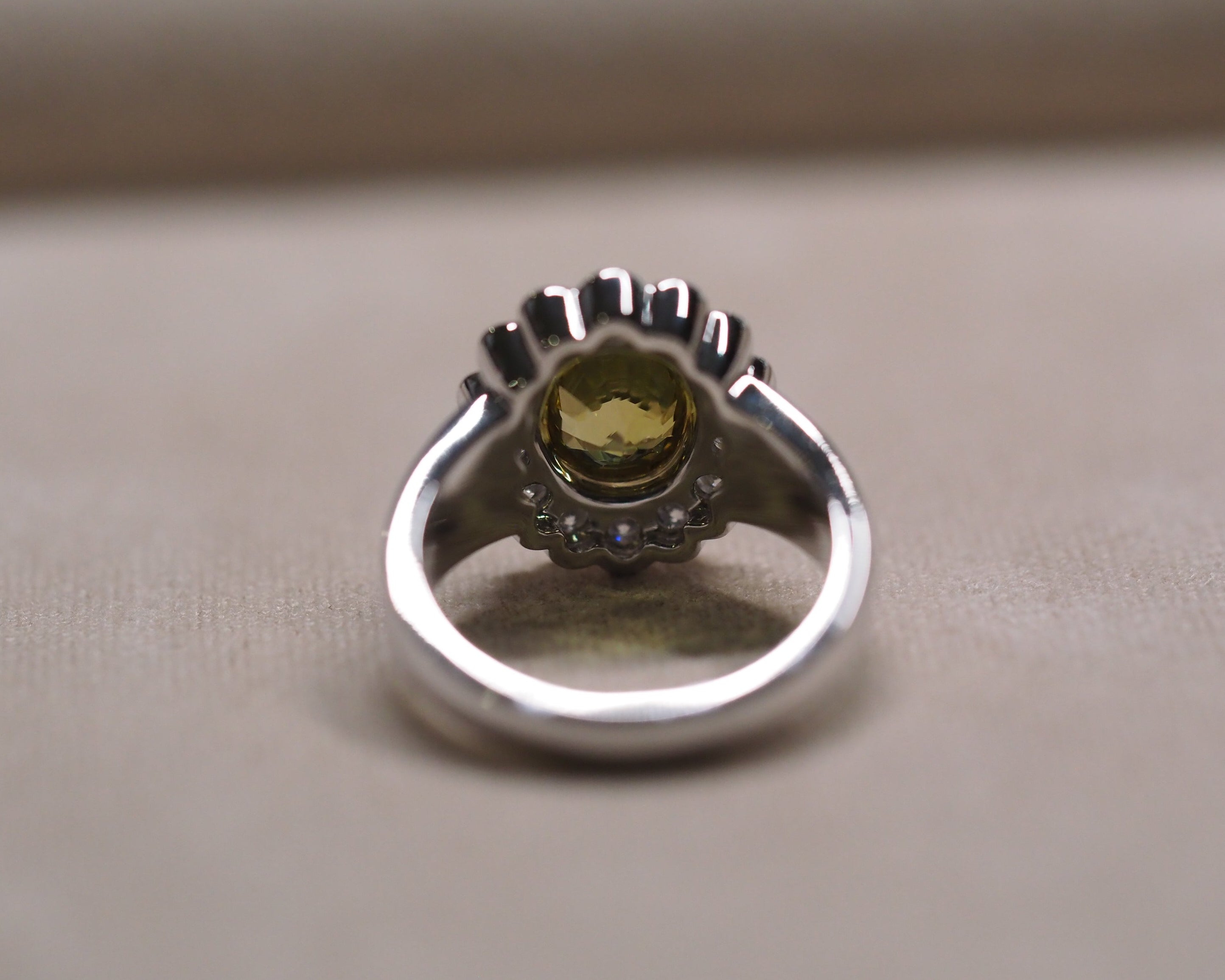 A silver ring featuring a central round yellow-green gemstone surrounded by small dark stones, set on a textured beige surface.