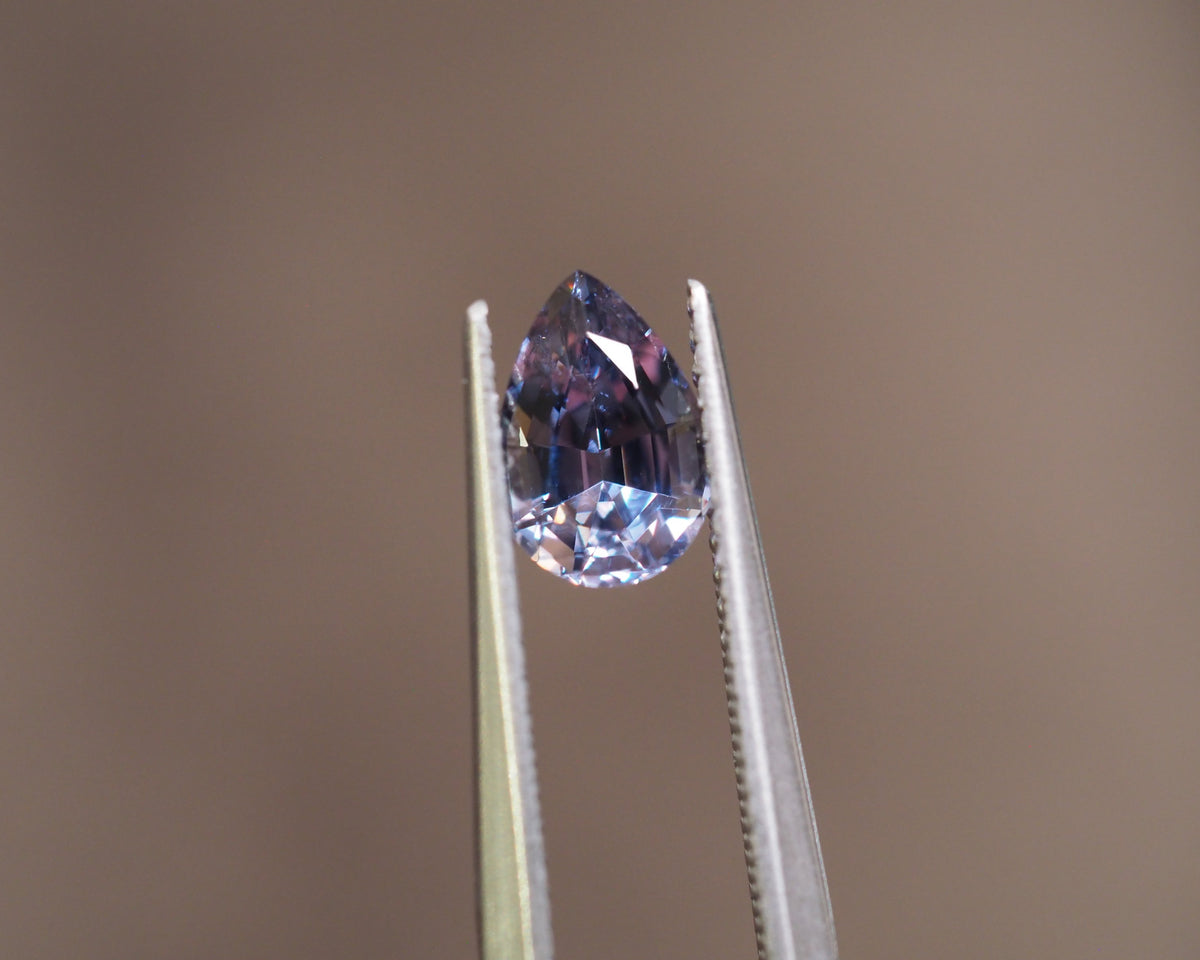 A pear-cut blue gemstone held in the jaws of metal tweezers against a solid warm brown background.