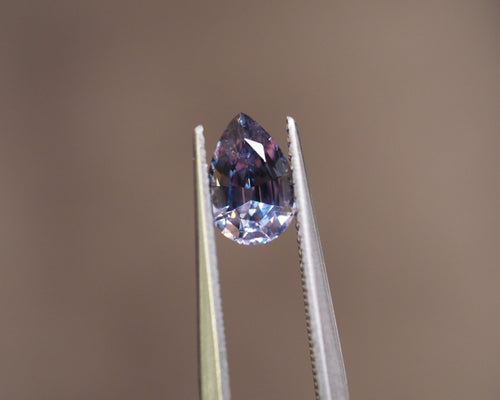 A pear-cut blue gemstone held in the jaws of metal tweezers against a solid warm brown background.
