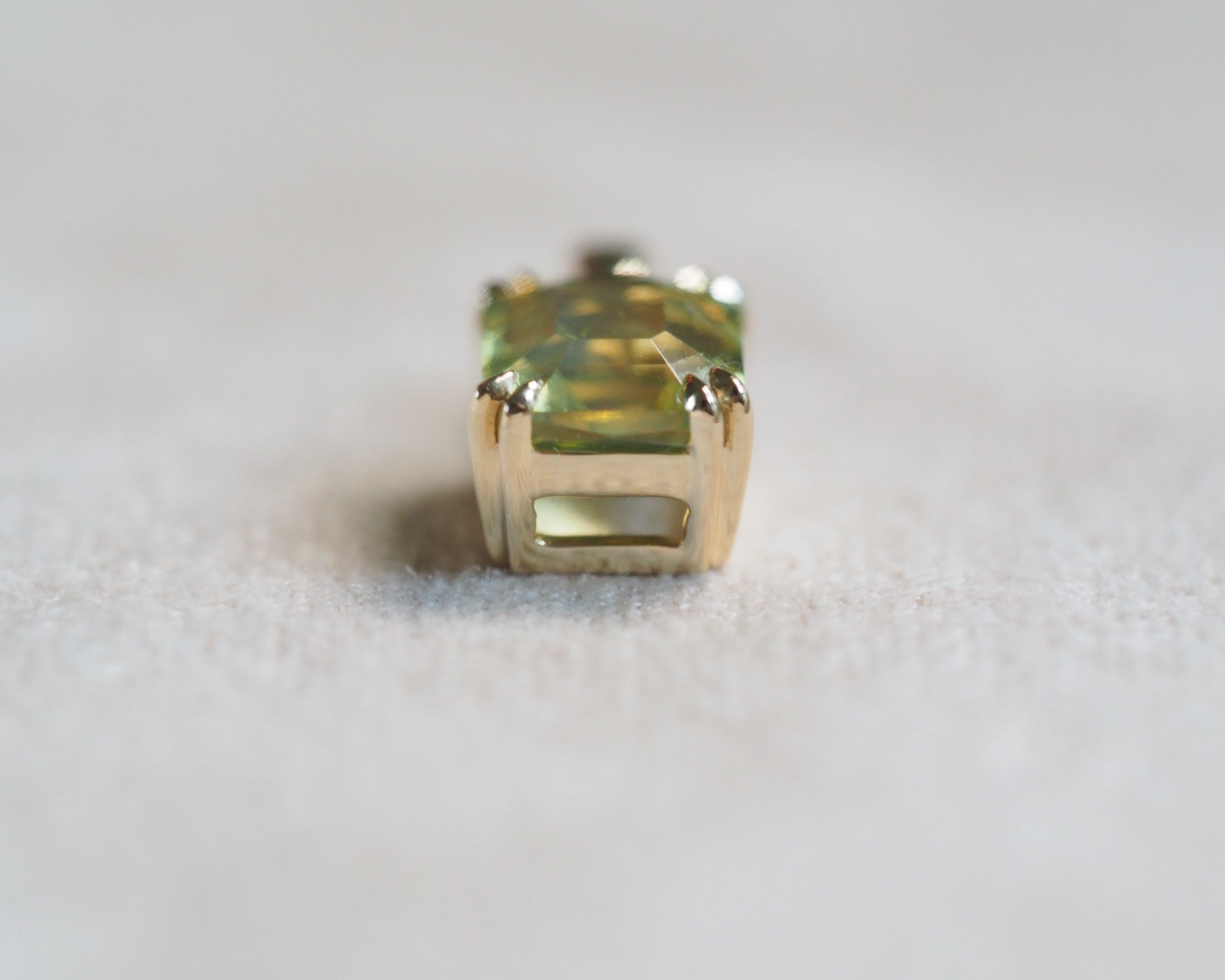 A close-up shot of a gold ring setting with a green rectangular gemstone, resting on a soft, light-colored textured surface.