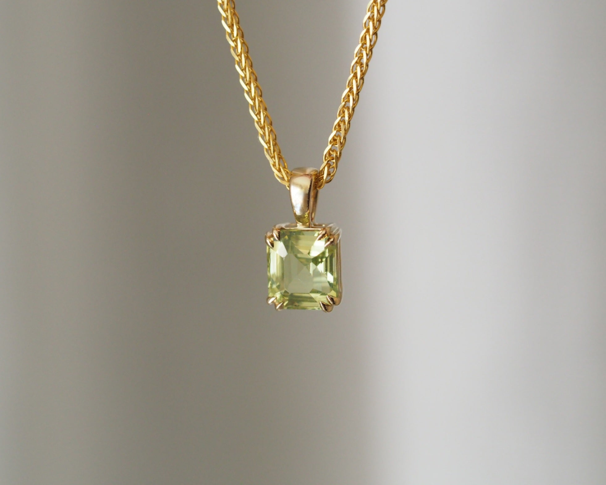 A gold chain necklace with a square-cut light green gemstone pendant, suspended against a soft gray gradient background.