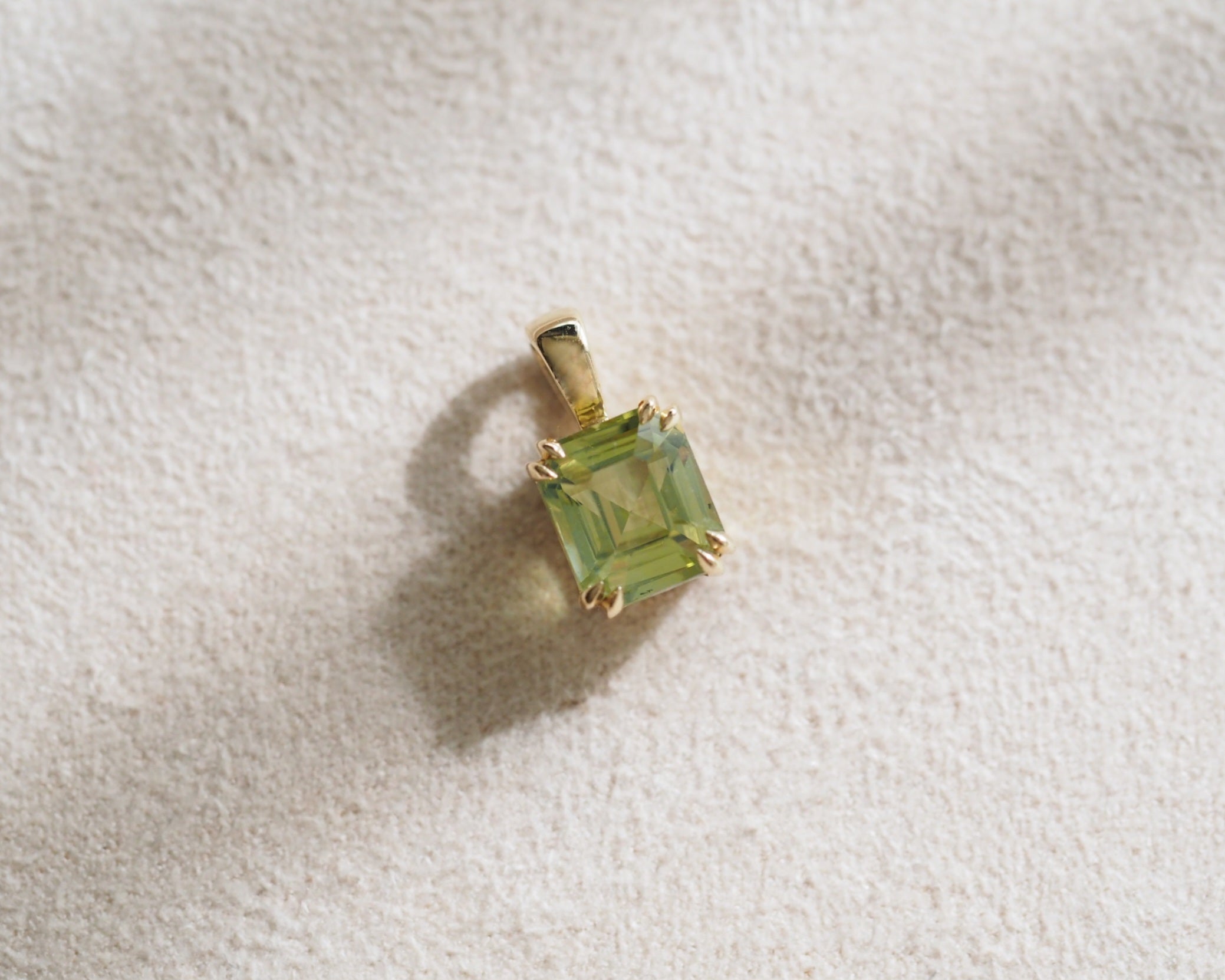 A assher-cut chrysoberyl gemstone pendant in a gold setting, resting on a textured off-white fabric surface.