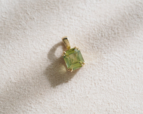 A assher-cut chrysoberyl gemstone pendant in a gold setting, resting on a textured off-white fabric surface.