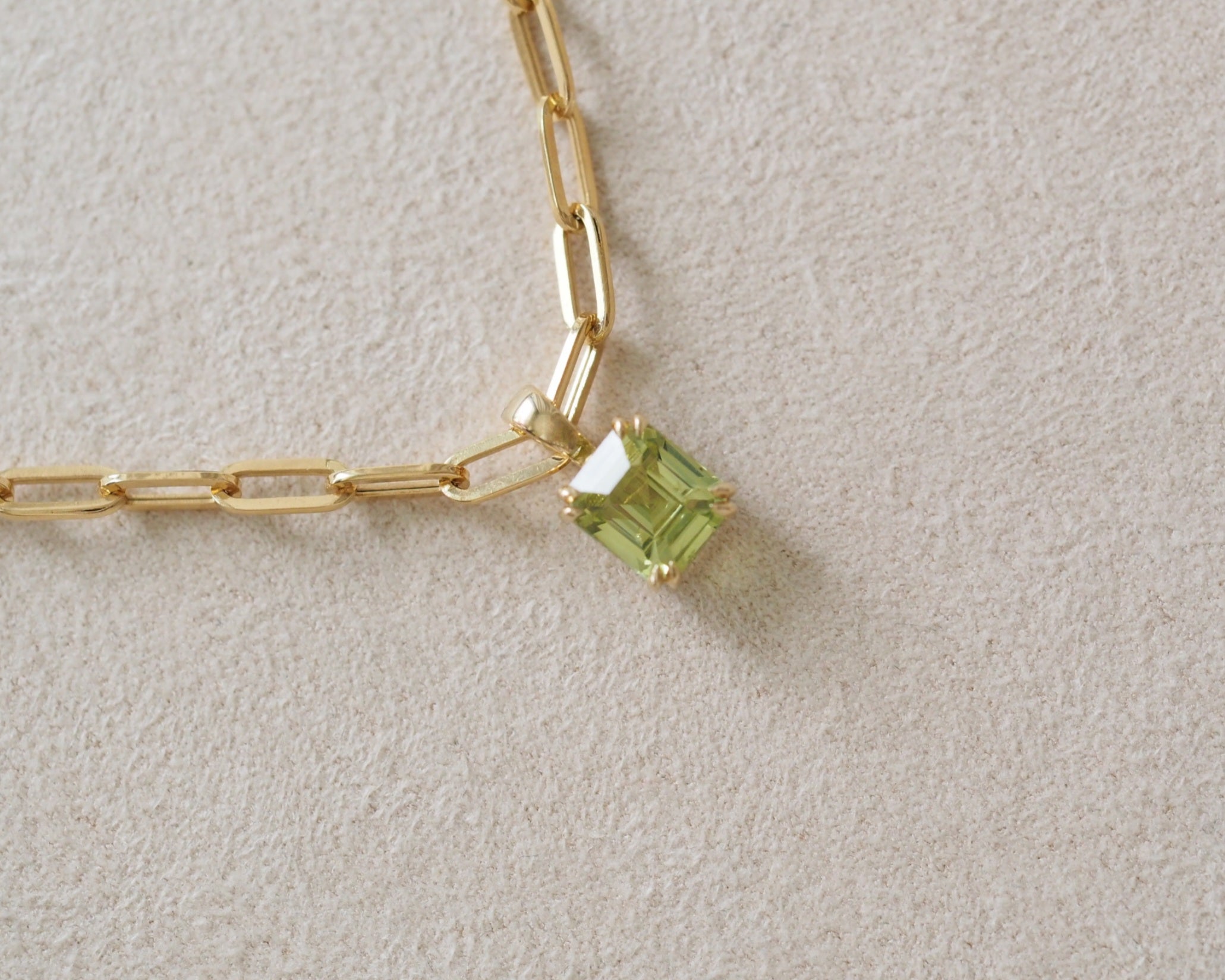 A close-up of a gold paperclip chain necklace with a small square-cut light green gemstone pendant, resting on a textured beige surface.