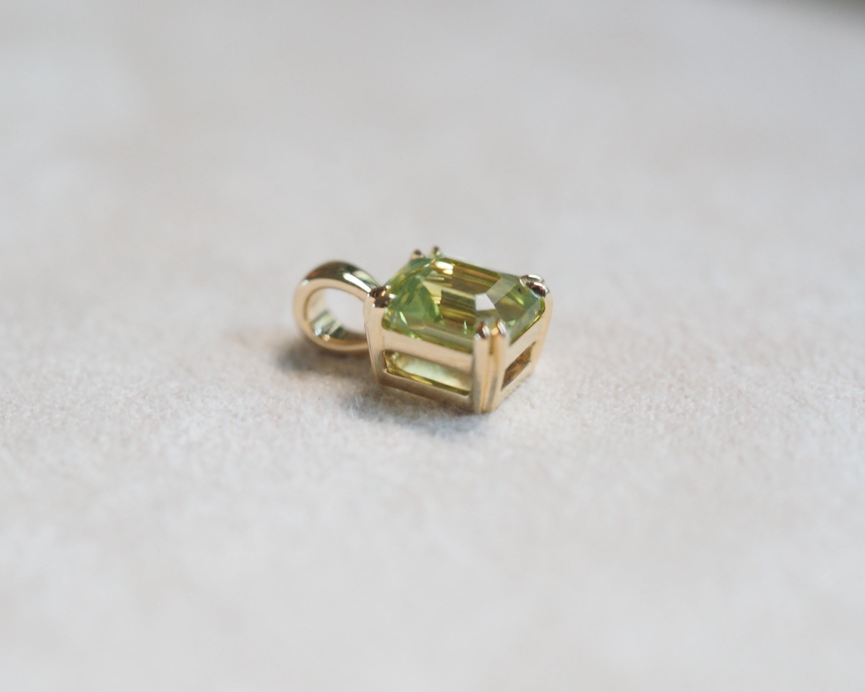 A small gold pendant with a rectangular green gemstone, photographed on a soft light gray textured surface.