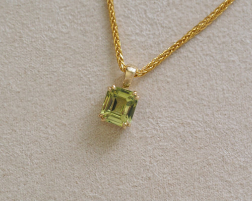 A gold chain necklace with a square-cut light green gemstone pendant, displayed on a textured light beige surface.