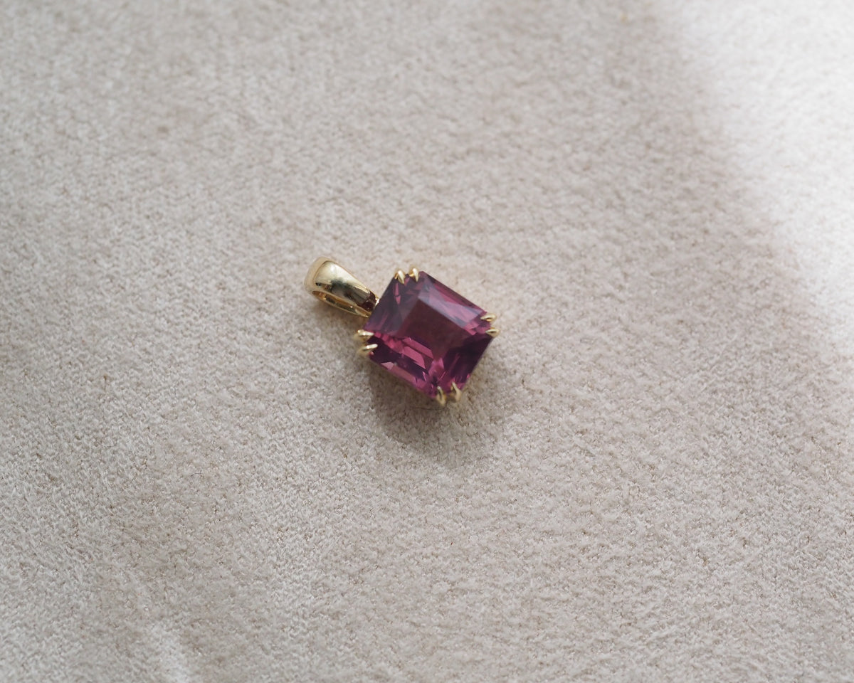 A small gold pendant with a rectangular faceted pink gemstone, resting on a textured beige fabric surface.