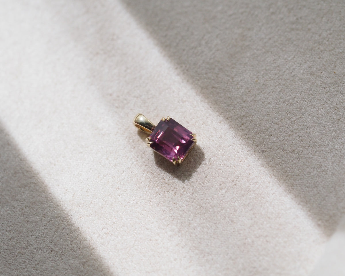 A small gold pendant with a square-cut pinkish-purple spinel, resting on a textured beige surface with soft diagonal shadows.