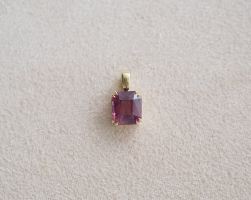 A rectangular pink spinel pendant set in a gold bezel, displayed on a textured light beige surface.