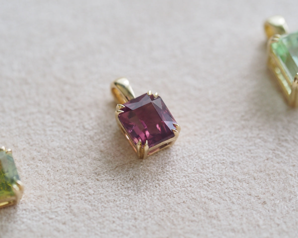 A close-up of a gold pendant with a cushion-cut pinkish-purple spinel, resting on a textured beige surface. Parts of other gold and green gemstone pendants are visible at the edges.