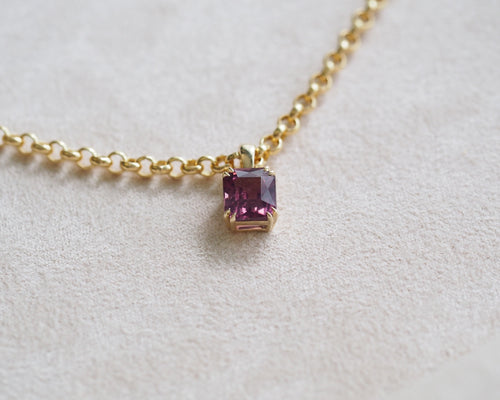 A gold chain necklace with a small square pinkish-purple spinel pendant, resting on a textured beige fabric background.