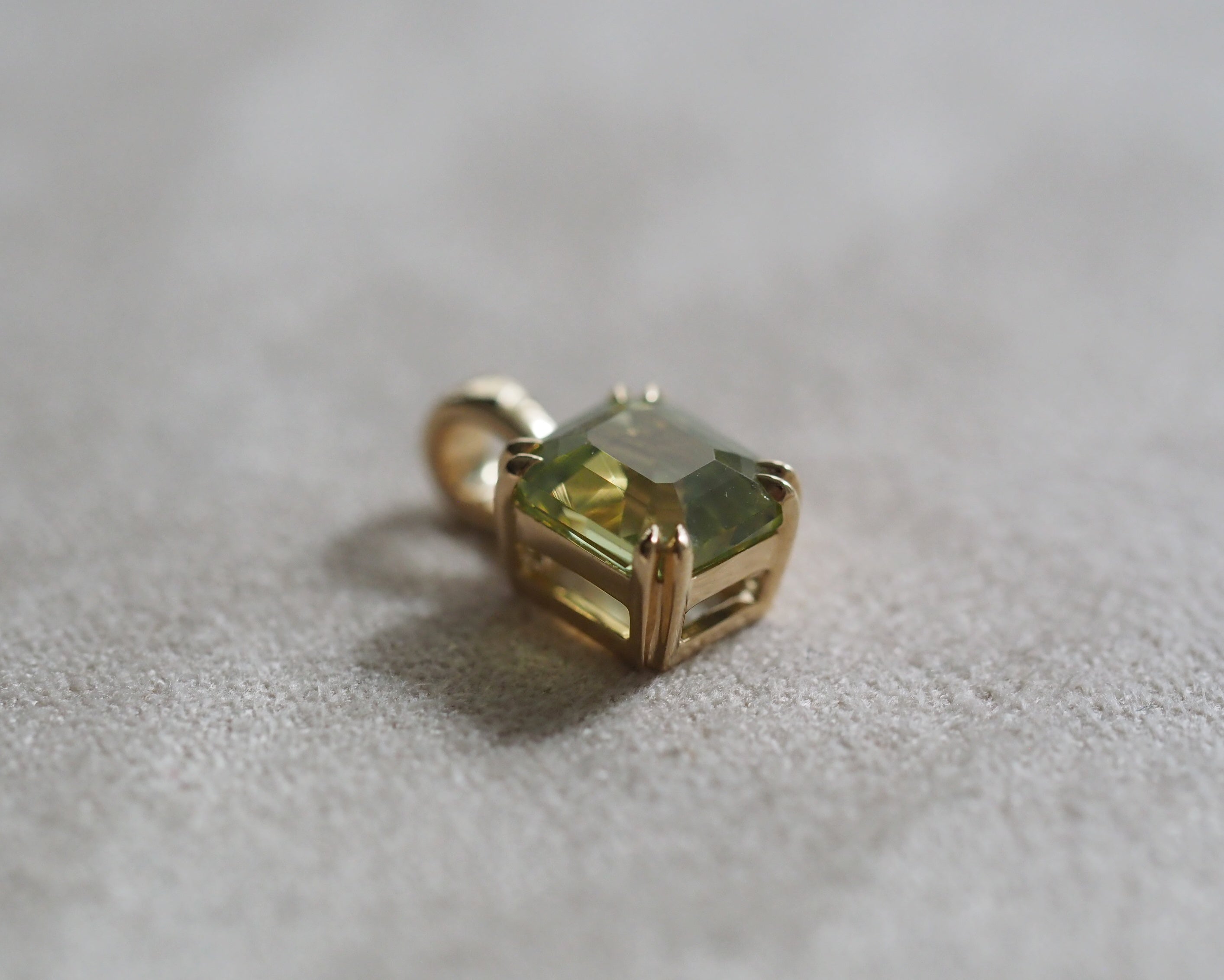 A gold pendant featuring a assher-cut lime green gemstone, resting on a textured light gray surface.