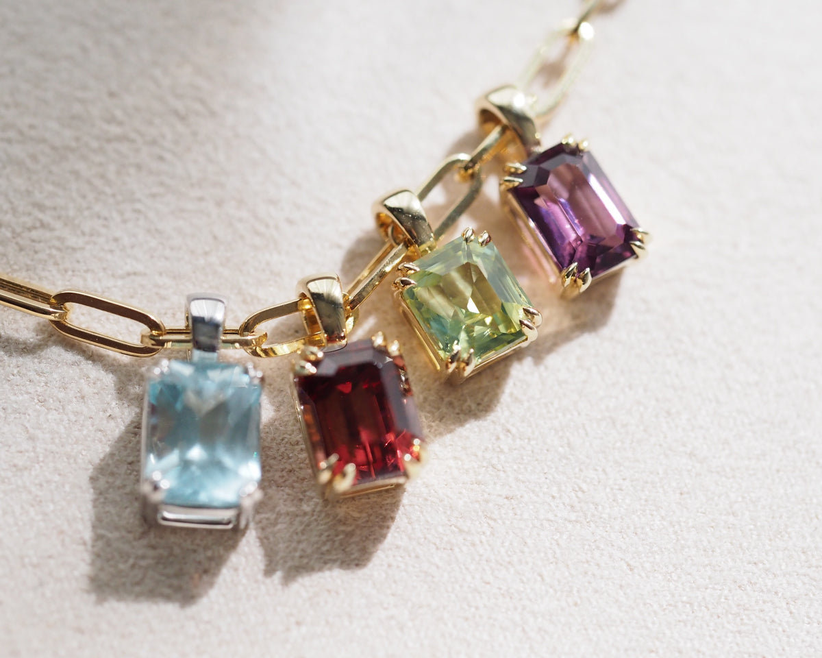 A close-up of a gold chain necklace with four rectangular gemstone pendants in different colors: light blue, dark red, light green, and purple, set on a textured light beige surface.