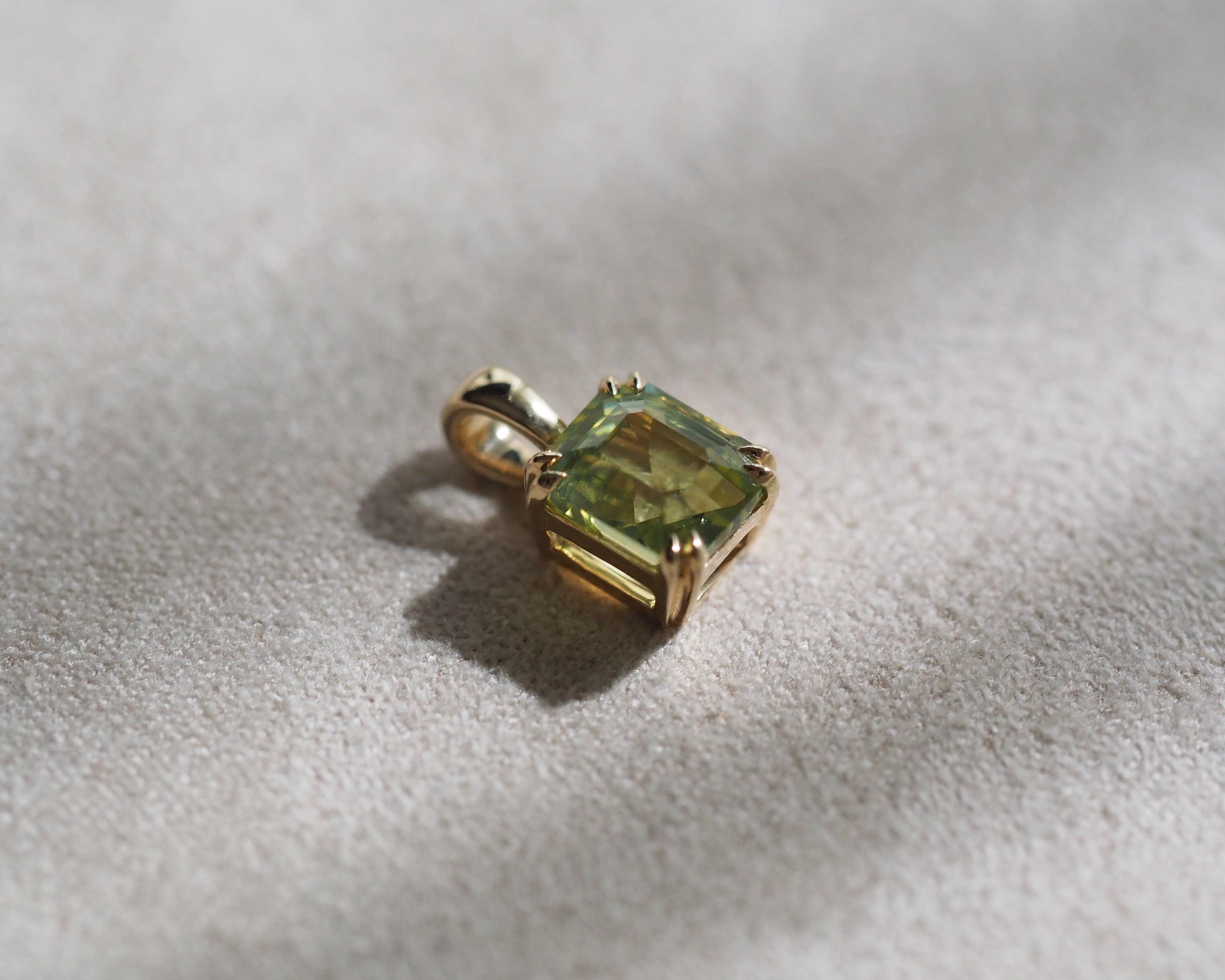 A gold pendant with a assher-cut light green chrysoberyl, resting on a textured beige fabric surface with soft shadows.