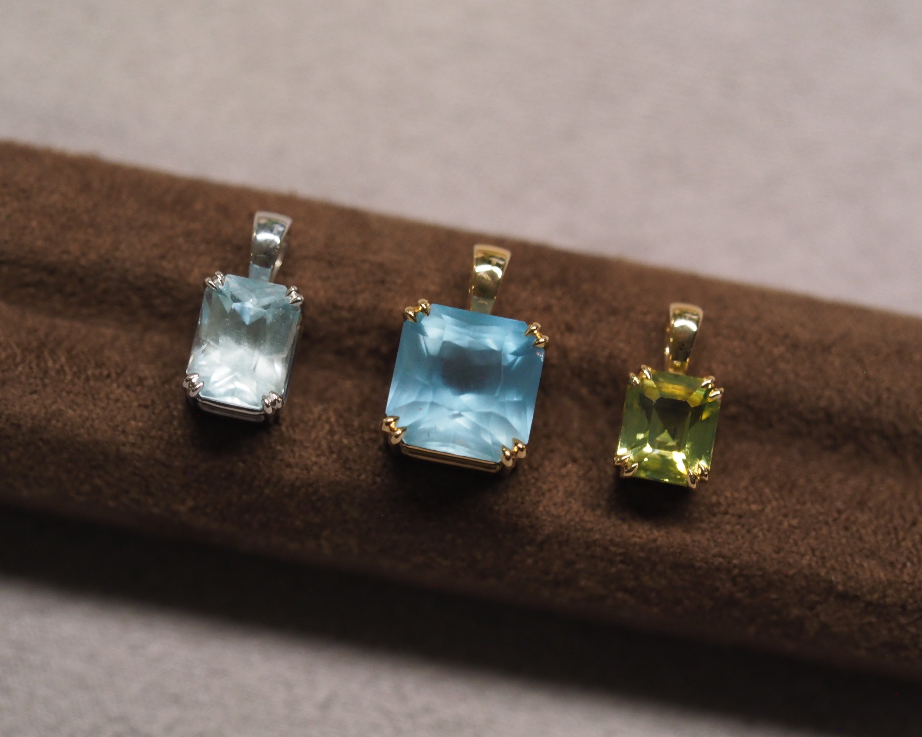 Three rectangular gemstone pendants displayed on a brown velvet jewelry roll. The center pendant features a light blue stone in a gold setting, flanked by a silver-toned pendant with a light blue stone on the left and a yellow-green stone on the right.