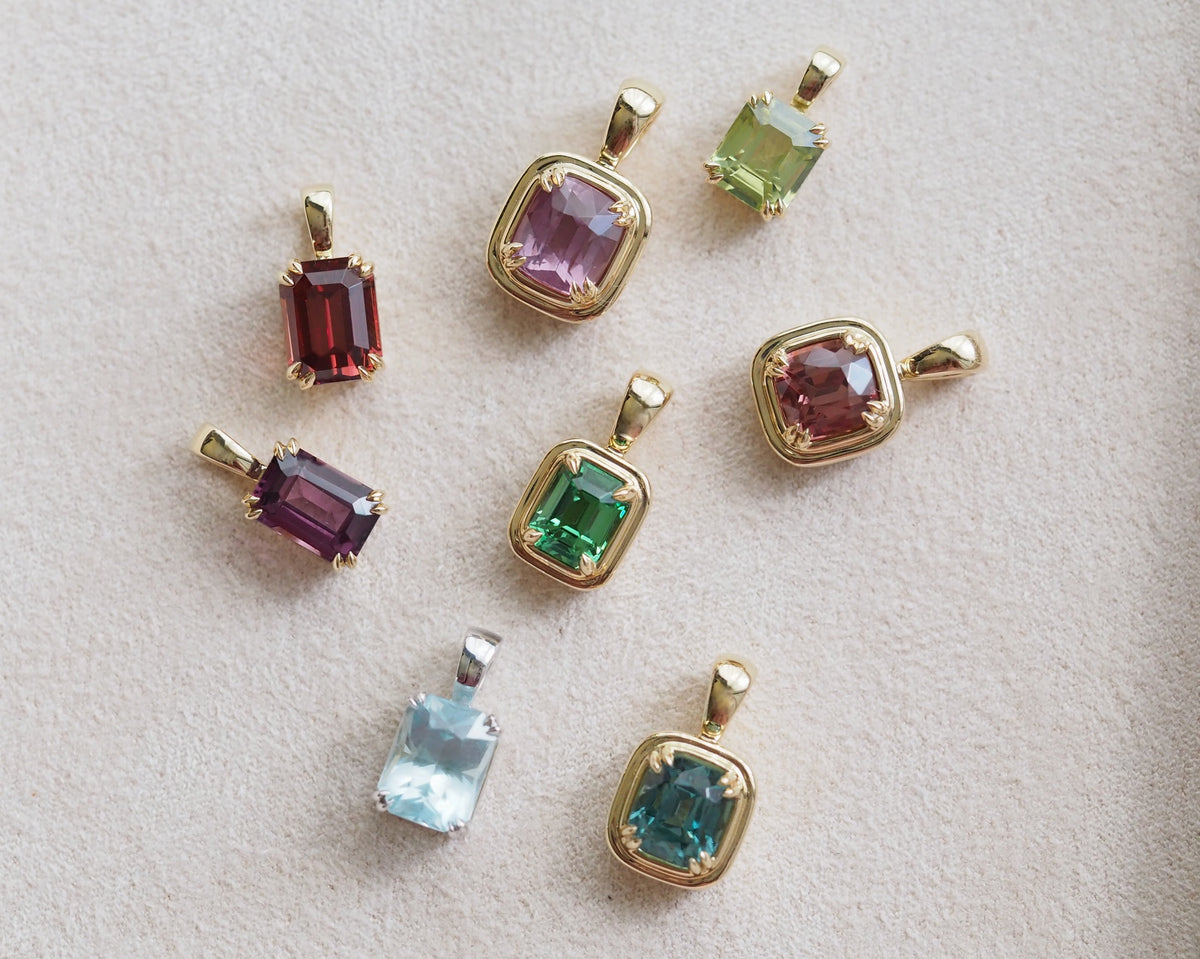 Seven gold pendant charms with different colored gemstones, including pink, green, red, purple, light blue, and teal, arranged on a textured light beige surface.