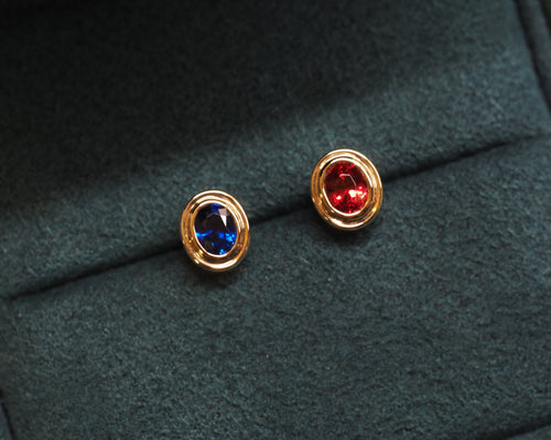 A pair of gold stud earrings with round blue and red gemstones, displayed on a dark green velvet jewelry box.