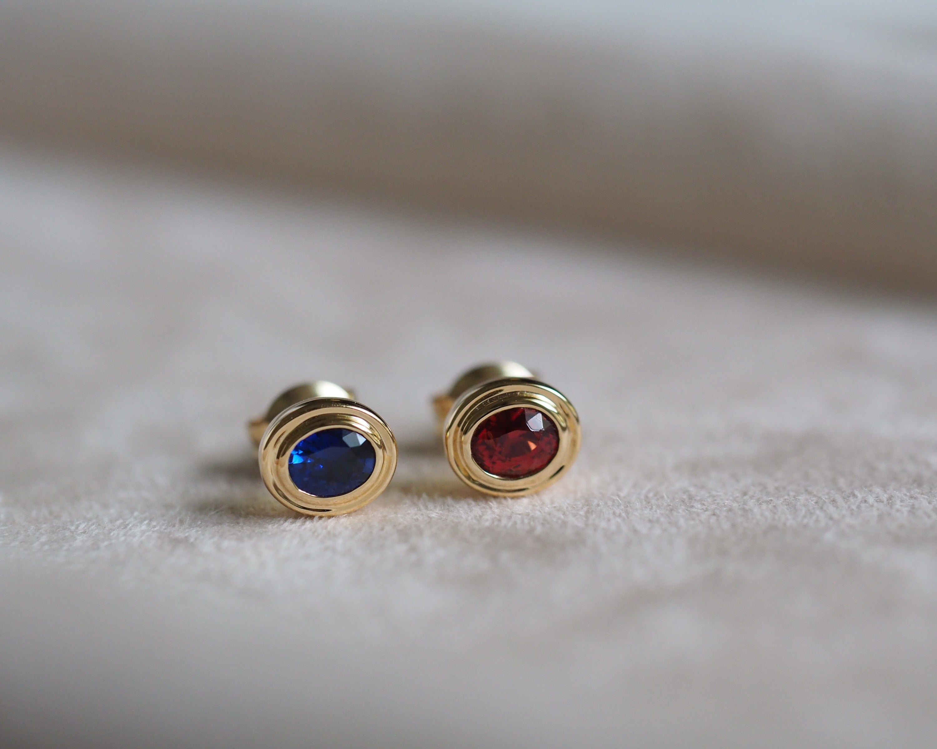 A pair of gold stud earrings with oval-cut blue and red gemstones, resting on a soft beige textured surface.