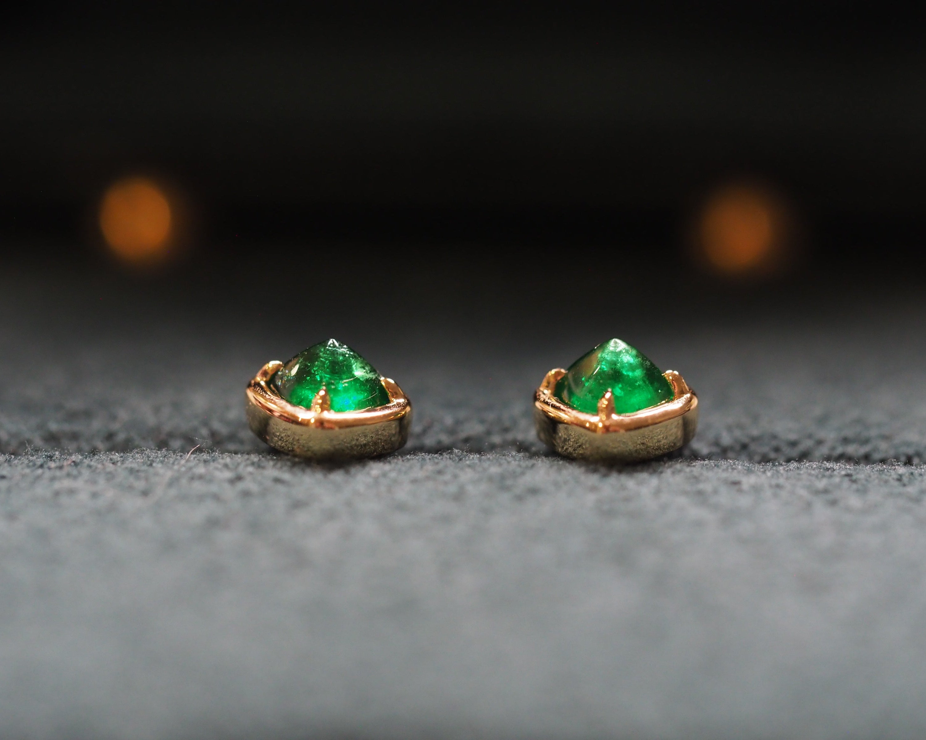 A pair of gold stud earrings with sugarloaf shape colombian emeralds, resting on a textured gray surface with a dark, blurred background.