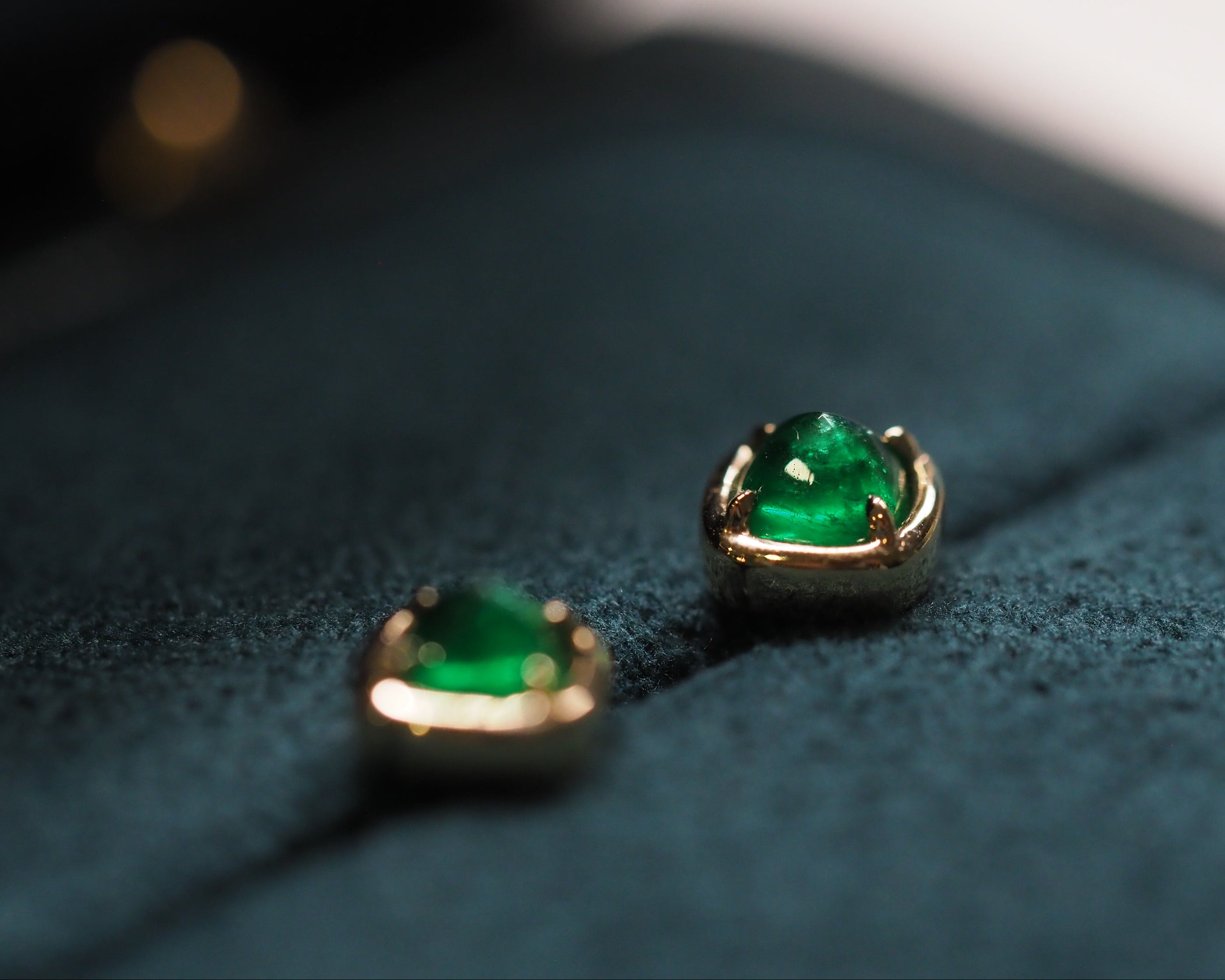 A pair of gold sugarloaf-shaped emerald earrings resting on a dark teal velvet jewelry box.