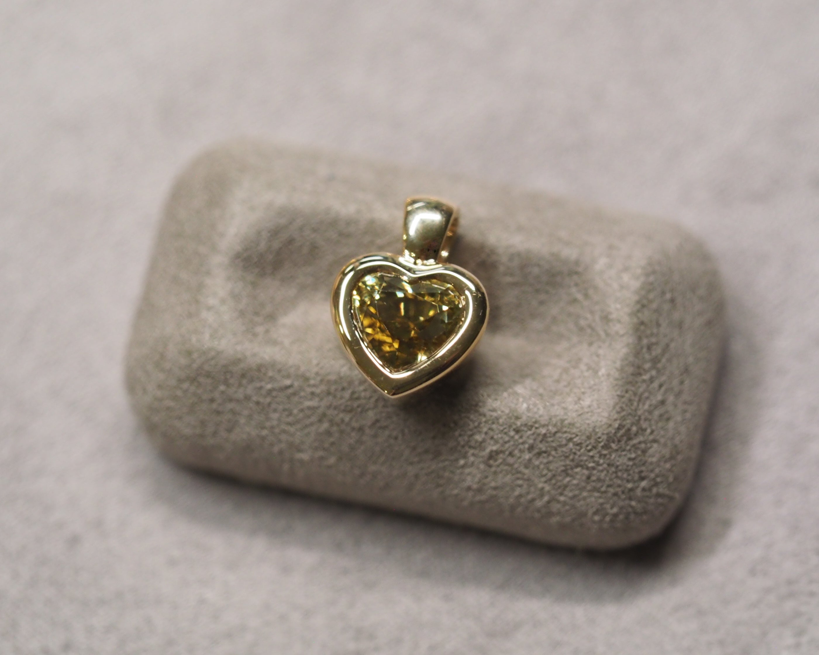 A small heart-shaped pendant with a yellow gemstone in a gold-toned setting, displayed on a textured gray stone stand against a soft gray background.
