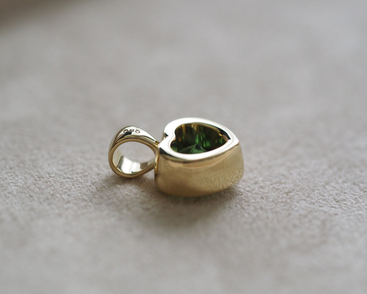 A small gold heart-shaped pendant charm with a green inlay, resting on a textured light gray surface.