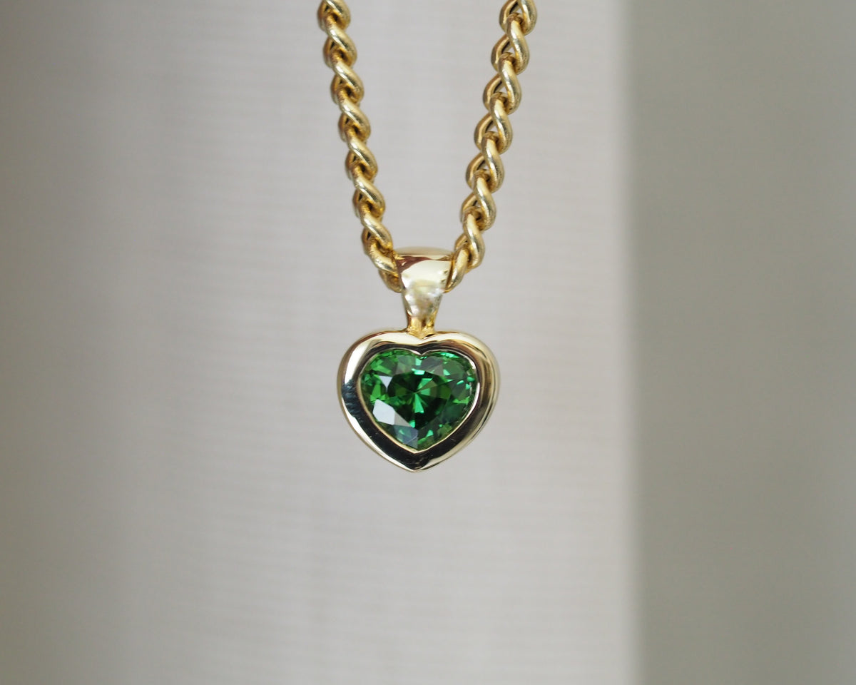 A gold heart-shaped pendant necklace with a faceted green gemstone, suspended from a twisted gold chain against a soft, light gray background.