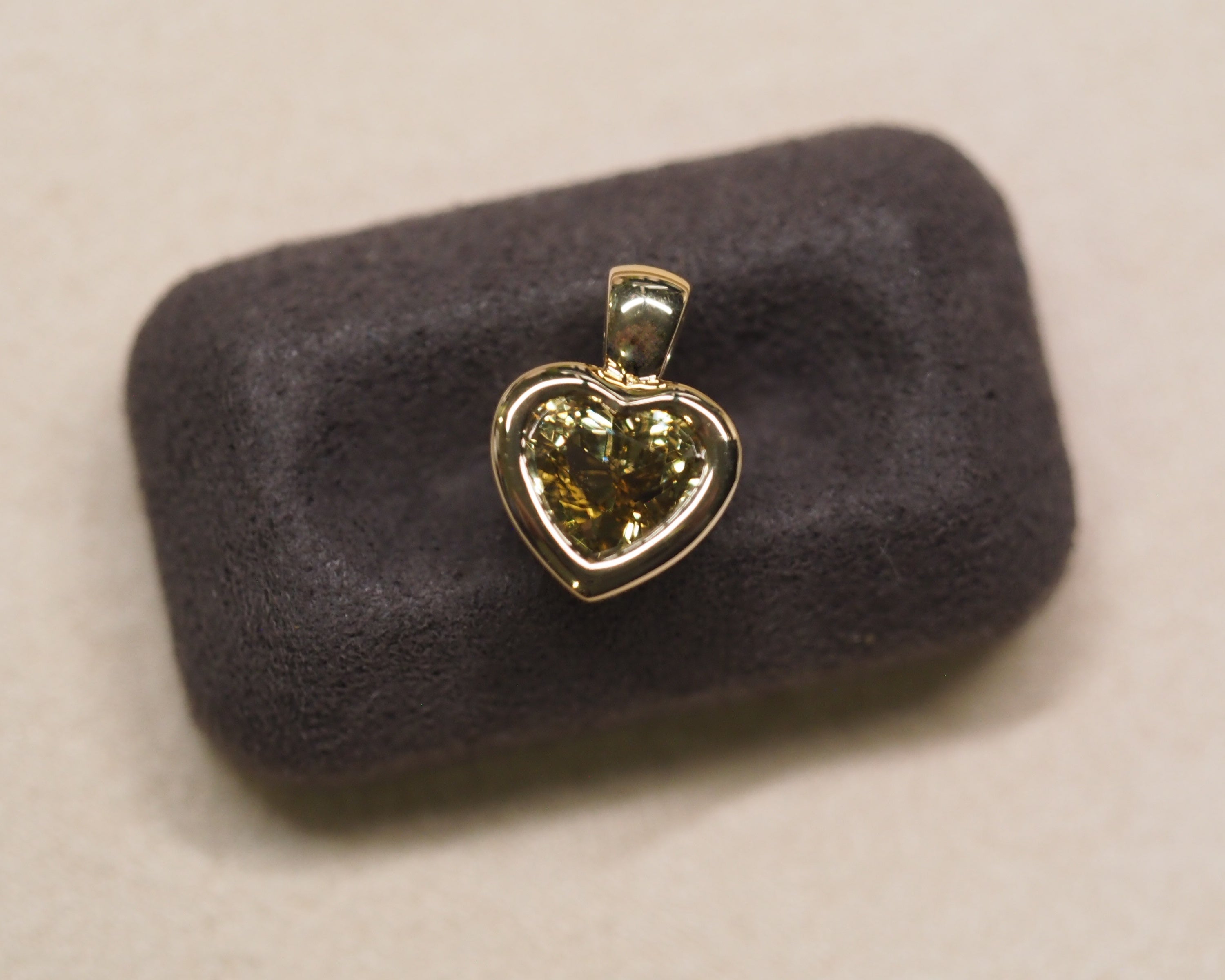 A heart-shaped yellow gemstone pendant in a gold setting, displayed on a dark gray velvet jewelry cushion against a light beige background.