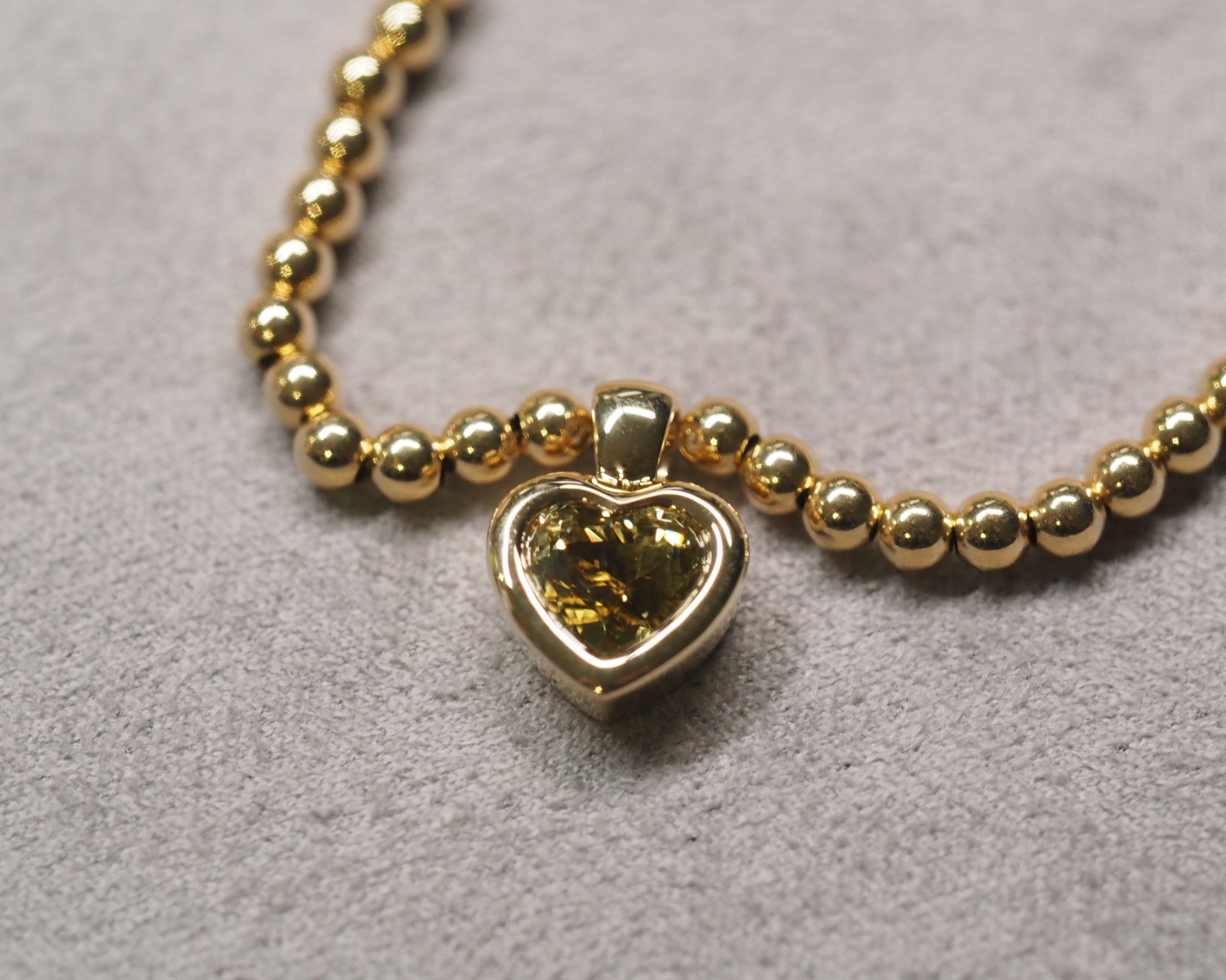 A gold heart-shaped pendant necklace with a yellow-green heart-shaped gemstone, resting on a textured gray surface.